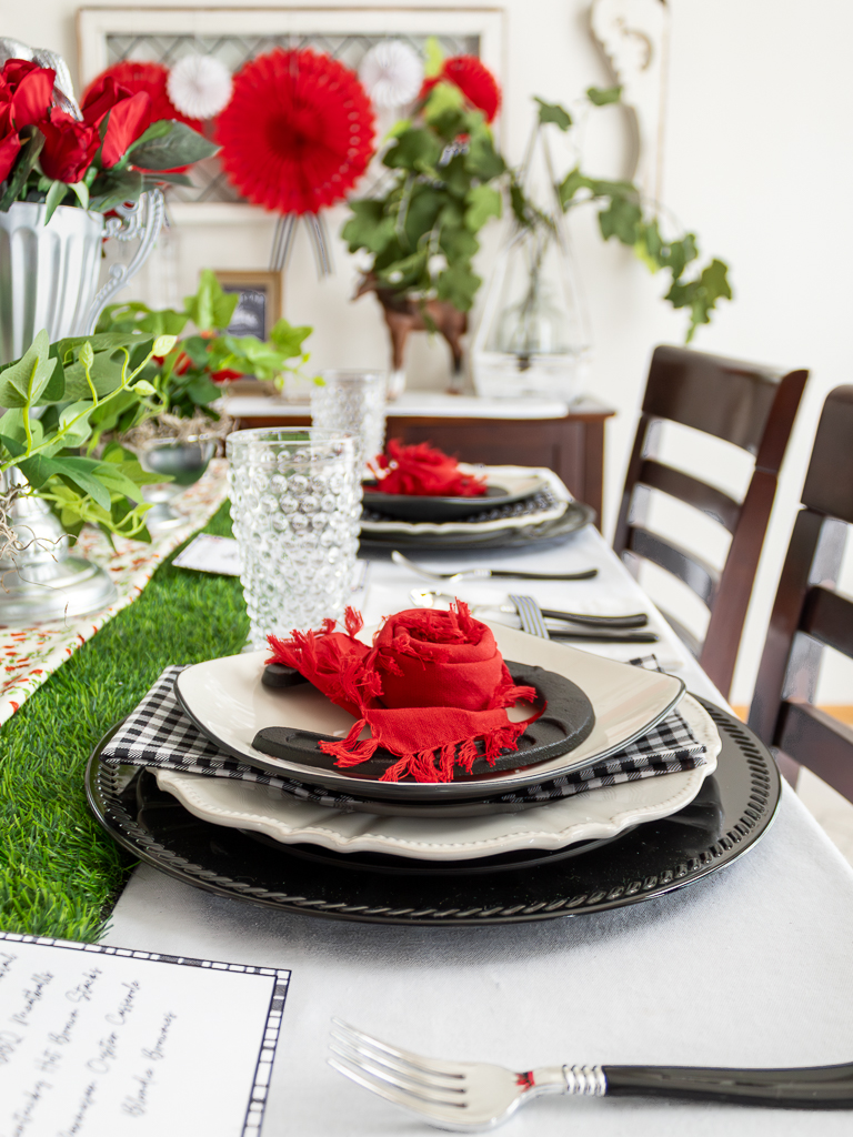 Close up view of a red and black place setting for a Kentucky Derby party tablescape with a black charger plate, gingham napkins, hobnail drinking glasses and a horsehoe and red napkin folded like a rose