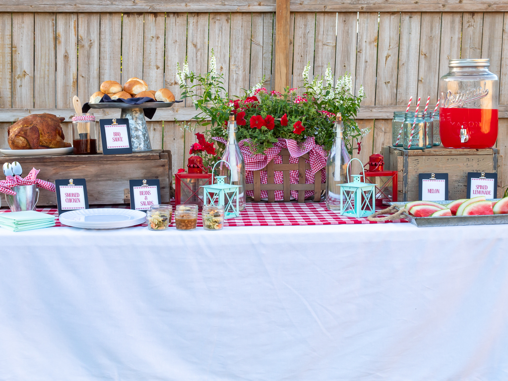 Food buffet table with red and white check tablecloth and picnic basket full of flowers set up for a backyard cookout