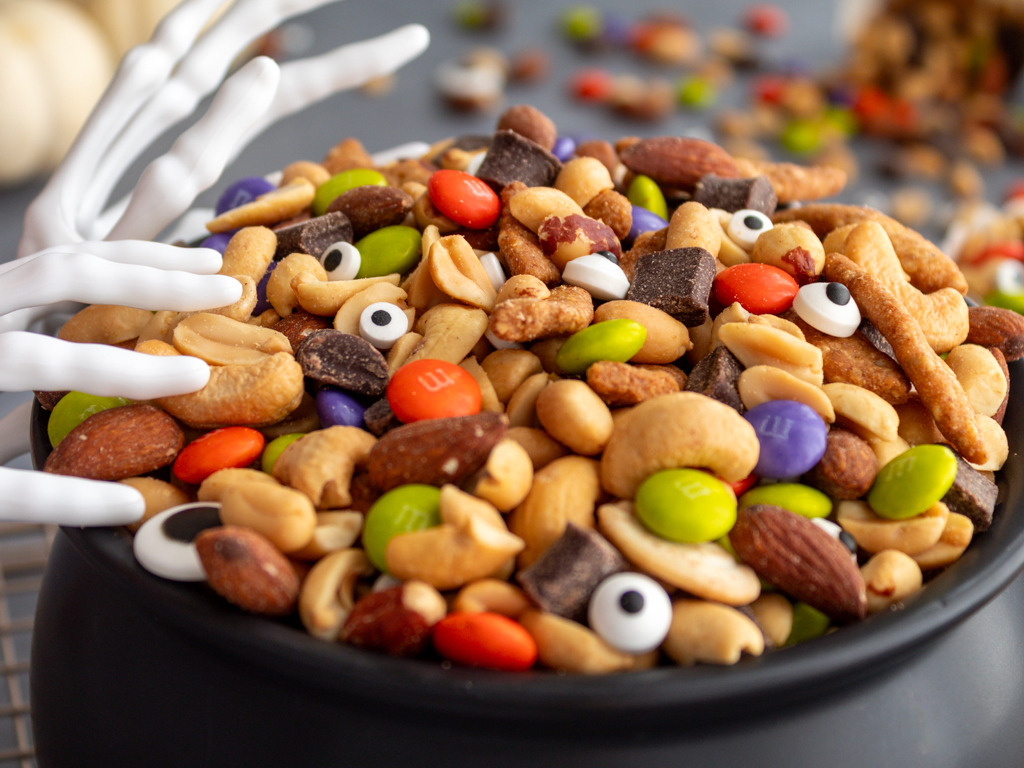 Close up of Halloween Monster Munch Trail Mix with candy eye sprinkles in a caldron bowl with spooky skeleton hand serving utensils on the side of the caldron