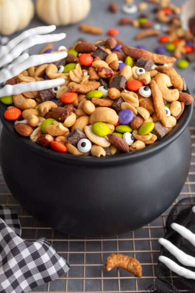 Halloween Monster Munch Trail Mix with candy eye sprinkles in a caldron bowl with spooky skeleton hand serving utensils on the side of the caldron and white pumpkins in the background