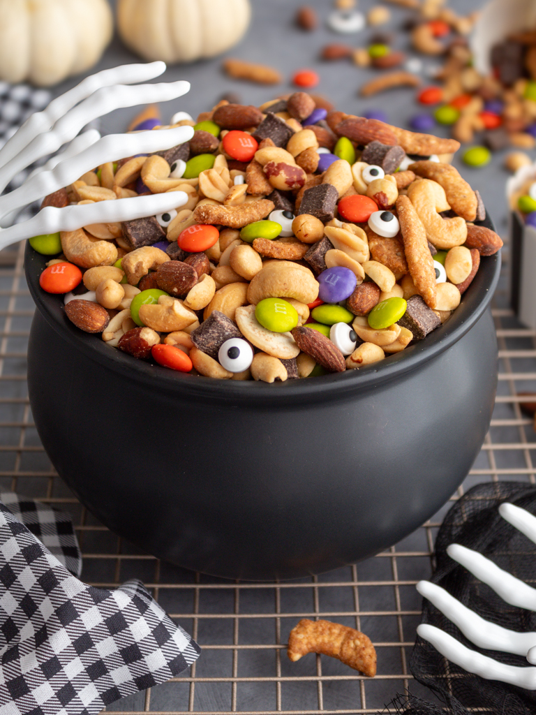 Halloween Monster Munch Trail Mix with candy eye sprinkles in a caldron bowl with spooky skeleton hand serving utensils on the side of the caldron and white pumpkins in the background