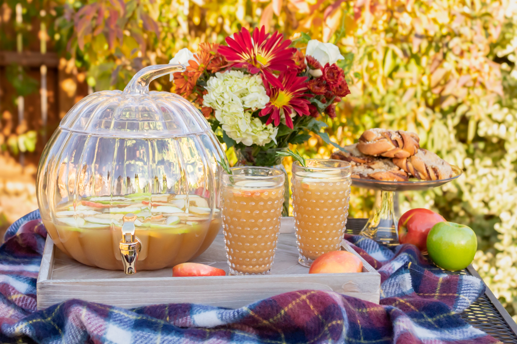 Two glasses of caramel aplple sangria  on a wooden serving tray with glass pumpkin drink dispenser and a cake plate with fall baked treats