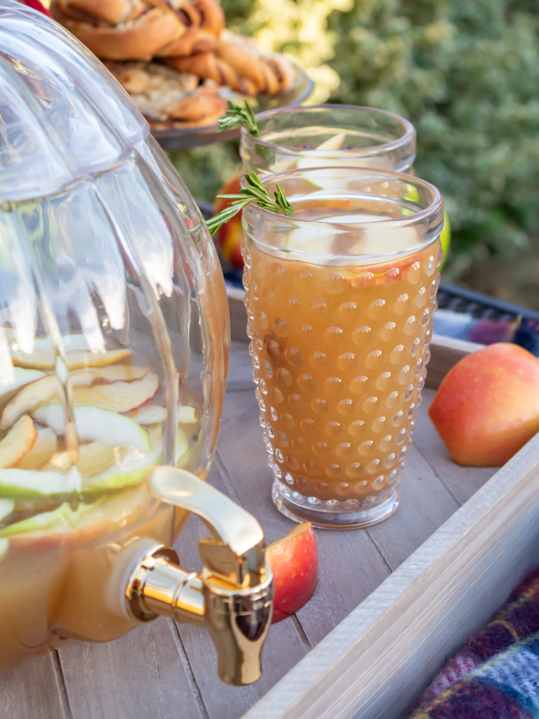 Two glasses of caramel aplple sangria  on a wooden serving tray with slices of apples and a glass pumpkin drink dispenser filled with more sangria