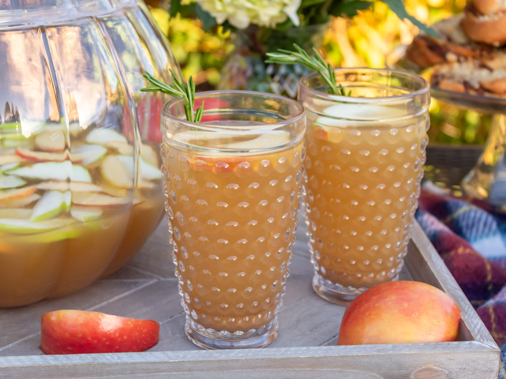 Two glasses of caramel aplple sangria  on a wooden serving tray with slices of apples and a glass pumpkin drink dispenser filled with more sangria