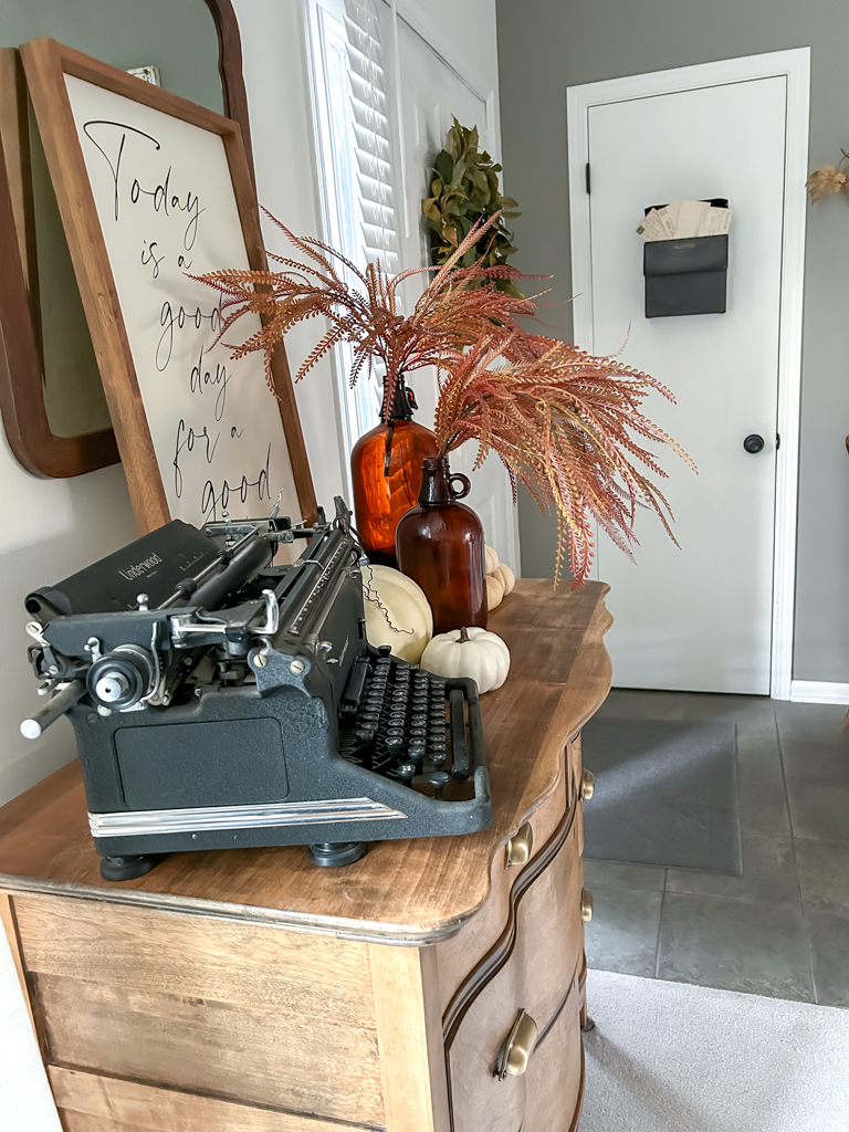 Fall vignette on an antique dresser wtih a vintage typewriter, pumpkins, and amber bottles with fall foliage