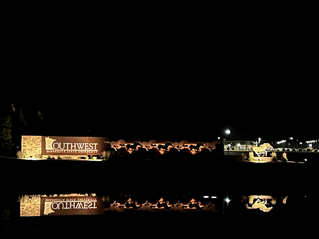 Main entrance to the SMSU campus wild mustang sign lit up a night with the reflection showing in the pond