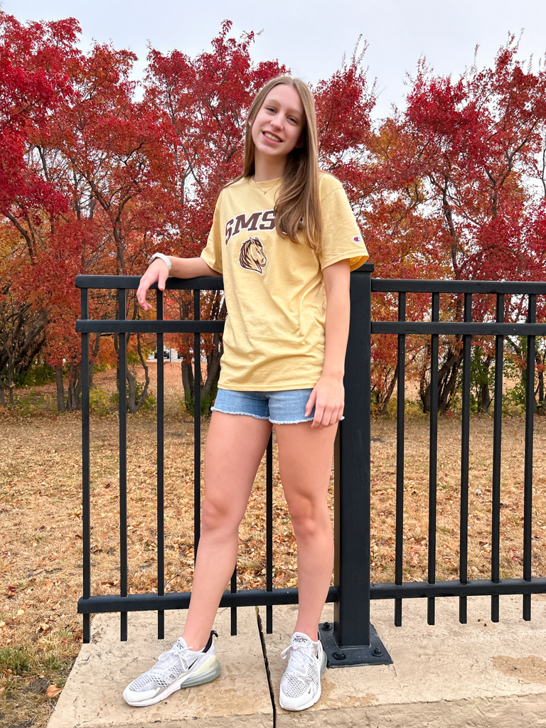 student posing in an SMSU shirt on campus in the fall with red leaves and trees in the background 