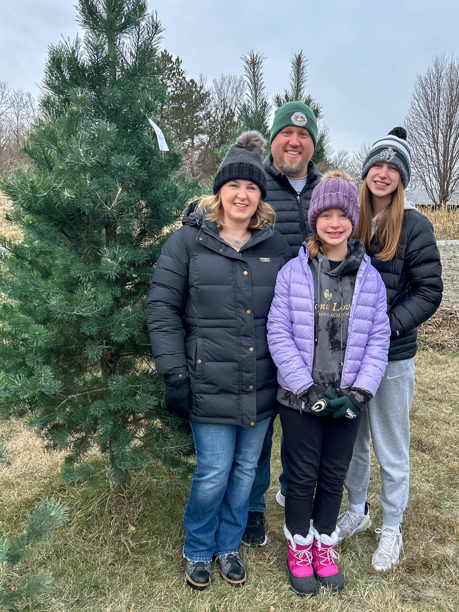 Family smiling in front of a reel tree they picked out for Christmas at a local tree farm