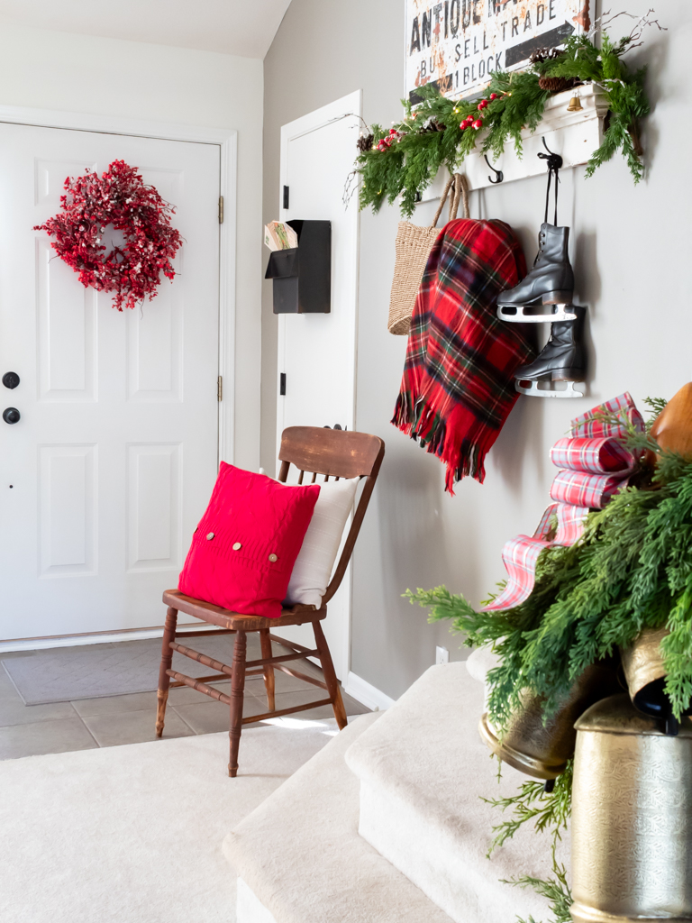 Front entry decorated for Christmas with a frosted red berry wreath hanging on the door, a shelf decorated with garland and twinkle lights haning on the wall with a plaid blanekt spilling out of a hanging basket, vintage black skates hanging from a hook on the shelf