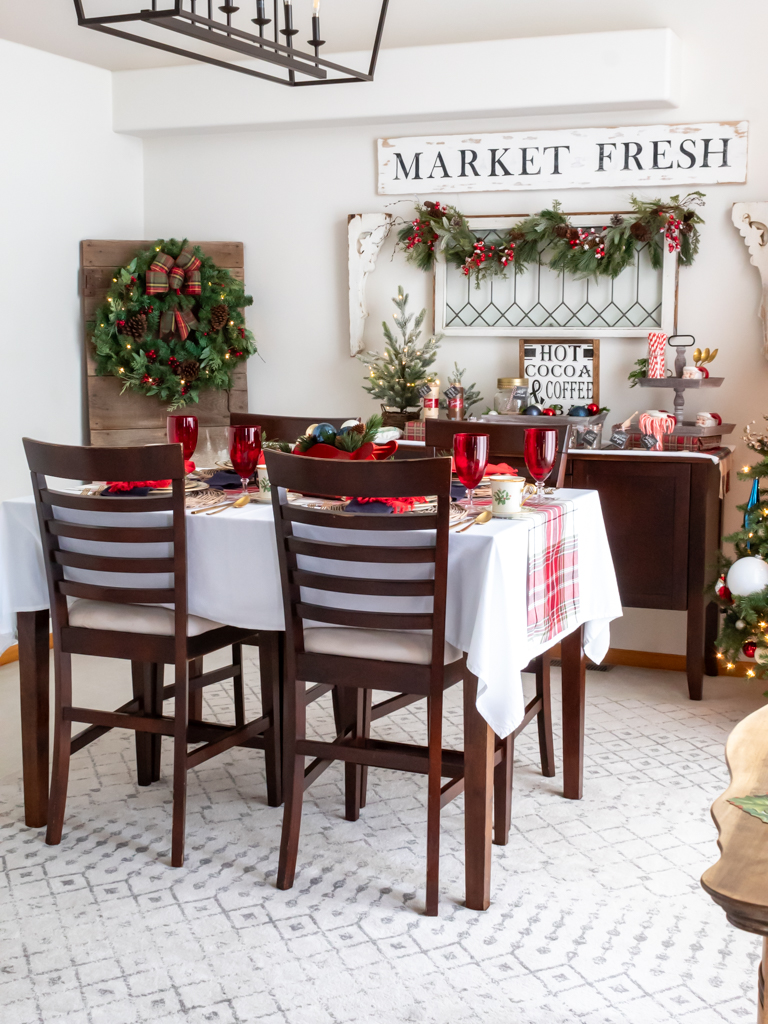 Dining room decorated for Christmas with red and blue decor, a tablescape set for a holiday meal, and a cozy hot cocoa bar set on a buffet in the background