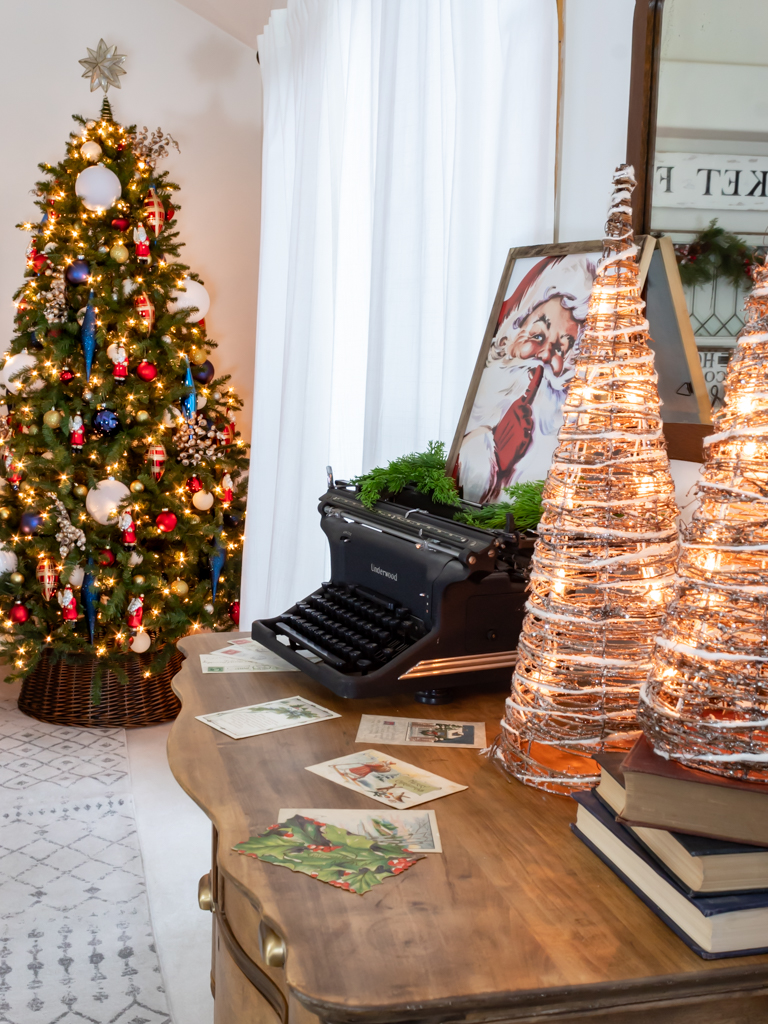 Vintage dresser with a christmas vignette including an old typewriter with a framed Santa print sitting on top, some light-up cone Christmas trees and a stack of red and blue vintage books with a large decorated Christmas tree in the background in the corner of the dining room