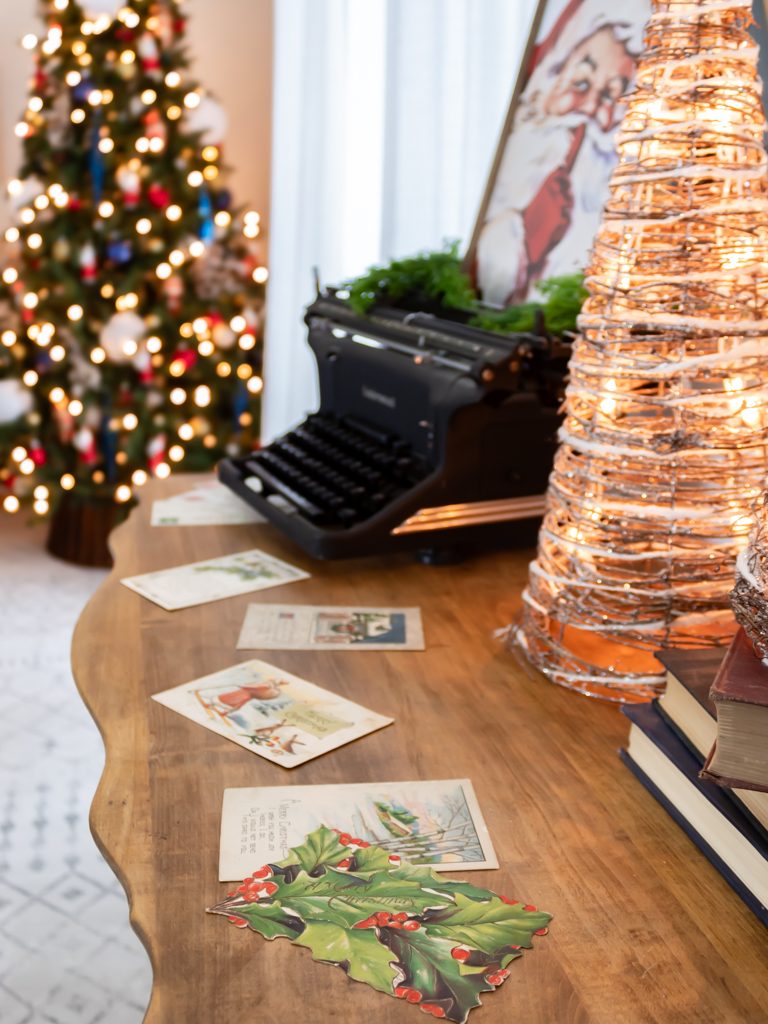 Vintage dresser in the dining room decorated with a christmas vignette including an old typewriter with a framed Santa print sitting on top, some light-up cone Christmas trees, scattered vintage postcards on top of the dresser and a large decorated Christmas tree in the background in the corner of the room