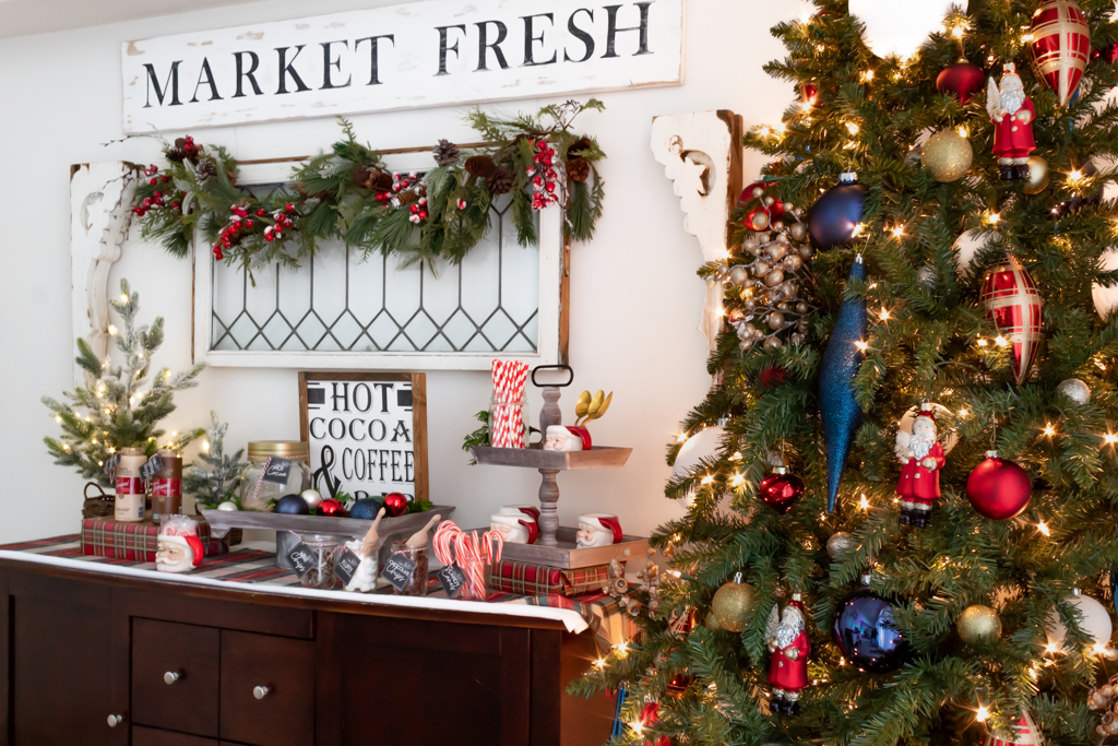 Cozy hot cocoa bar set on a buffet with a Christmas tree in the corner, and a vintage window above the cocoa bar draped with garland