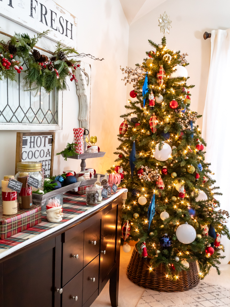 Large Christmas tree in the corner of the dining room decorated with red, white, and blue ornaments with a woven tree collar and a hot cocoa bar set up on a buffet in the foreground with a vintage leaded window hanging above it on the wall with a garland draped on it