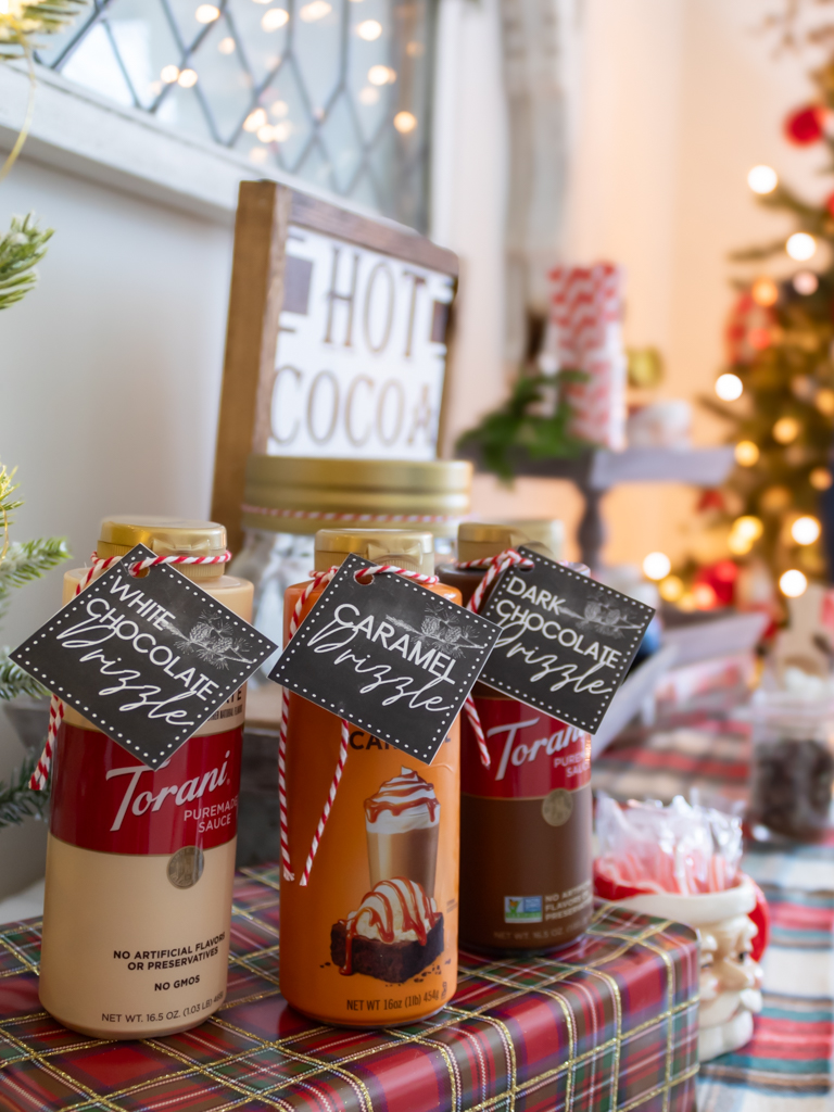 A hot cocoa bar with bottles of caramel and chocoalte sauce with  free printable hot cocoa bar tags tied on and a hot cocoa sign and tiered tray with santa mugs in the background