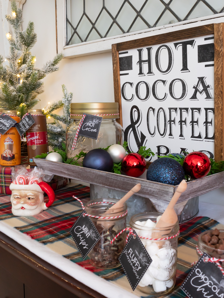 Hot cocoa sign styled on a tiered tray with red and blue Christmas ornaments on a cocoa bar with other decor including a vintage santa mug filled with mini candy canes and small bar jars with wooden scoops, filled with hot cocoa toppings with tied on free printable hot cocoa bar tags 