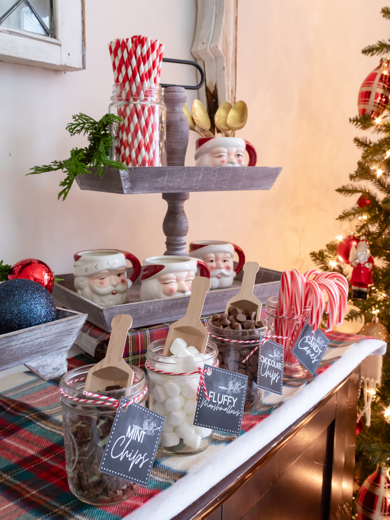 Tiered tray on a cocoa bar with  a jar filled with red and white paper straws, Santa Christmas mugs, and small bar jars with wooden scoops, filled with hot cocoa toppings with free printable hot cocoa bar tags tied on