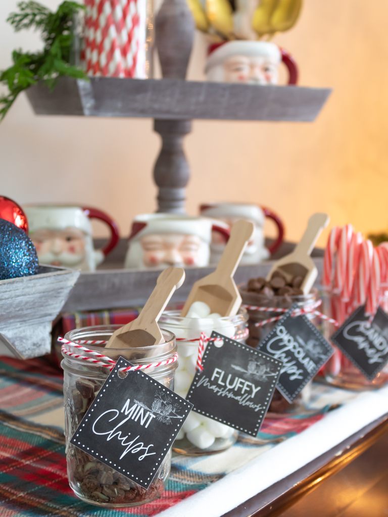 A hot cocoa bar with small bar jars with wooden scoops, filled with hot cocoa toppings with  free printable hot cocoa bar tags tied on and a tiered tray with santa mugs in the background