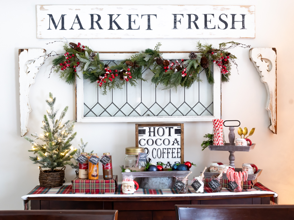 Cozy hot cocoa bar set on a buffet with plaid and red holiday decorations and a vintage window hanging on the wall above it draped with a Christmas garland