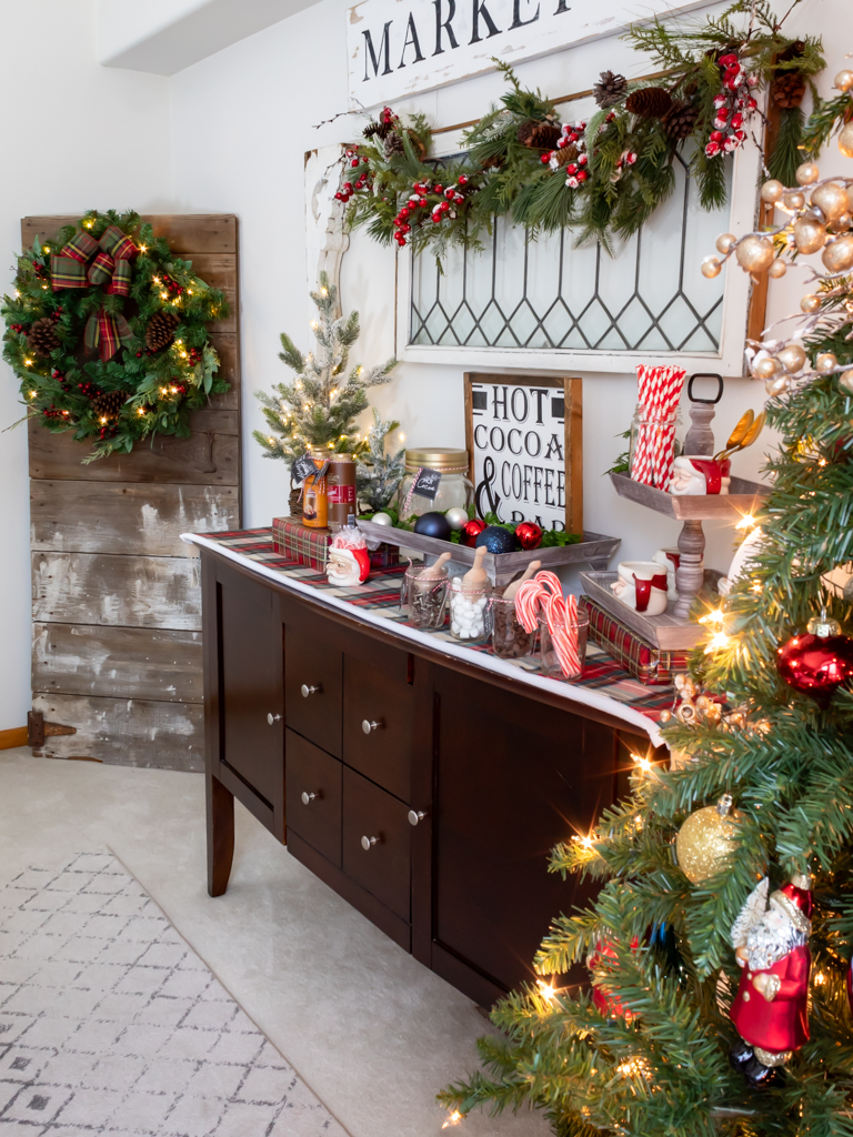 Cozy hot cocoa bar set on a buffet with plaid and red holiday decorations, a vintage window hanging on the wall above it draped with a Christmas garland and a barn door propped up in the  corner of the room with a large holiday wreath hanging on it