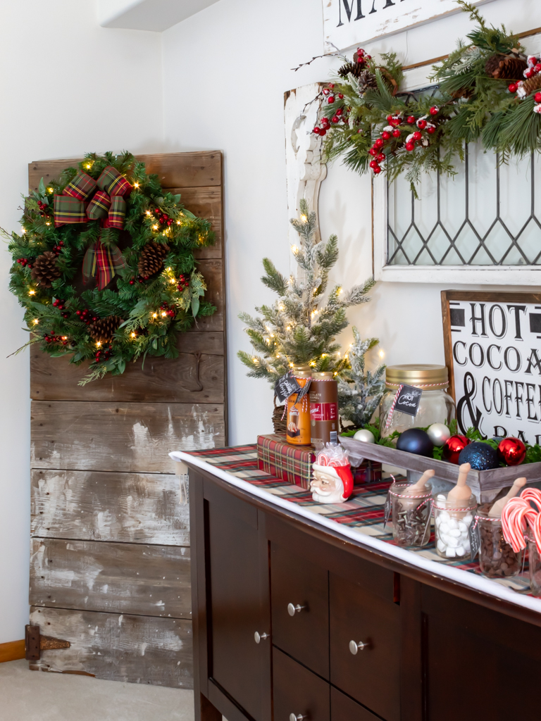 Cozy hot cocoa bar set on a buffet with plaid and red holiday decorations, a vintage window hanging on the wall above it draped with a Christmas garland and a barn door propped up in the  corner of the room with a large holiday wreath hanging on it