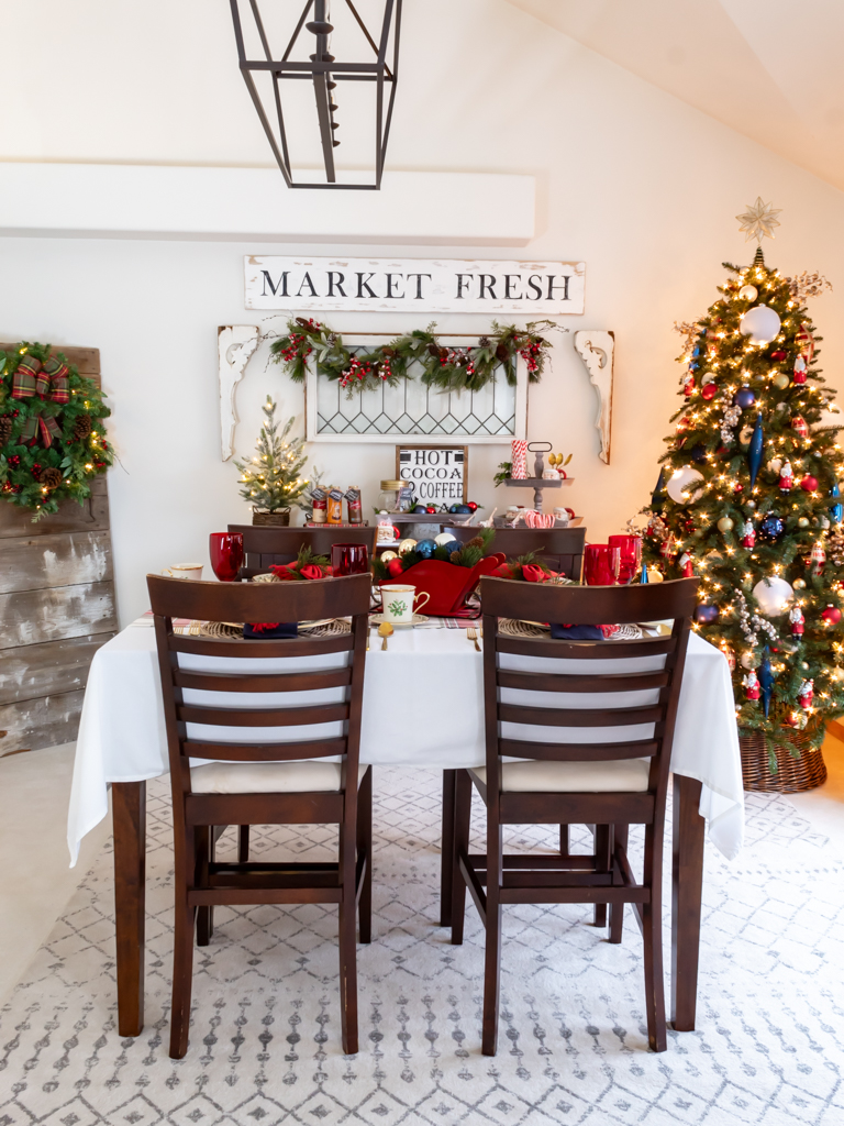 Dining room decorated for Christmas with red and blue decor, a tablescape set for a holiday meal, and a cozy hot cocoa bar set on a buffet in the background and a decorated Christmas tree in the corner