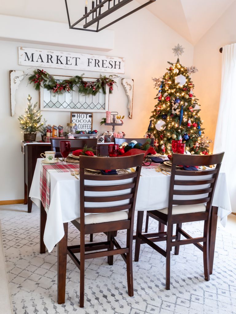 A tablescape set for a holiday meal, with red and blue decorations including Lenox Christmas china, a red fringed napkin with a greenery napkin ring, red goblets, a red sleigh centerpiece filled with ornaments a decorated Christmas tree in the corner of the room, and a buffet decoratd with holiday greenery adn a cozy hot cooca bar