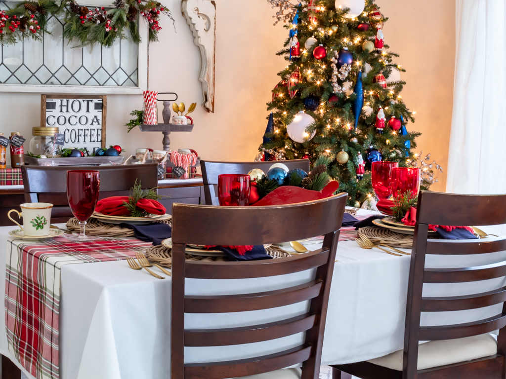 Dining room decorated for Christmas with red and blue decor, a tablescape set for a holiday meal, and a cozy hot cocoa bar set on a buffet in the background and a Christmas tree in the corner