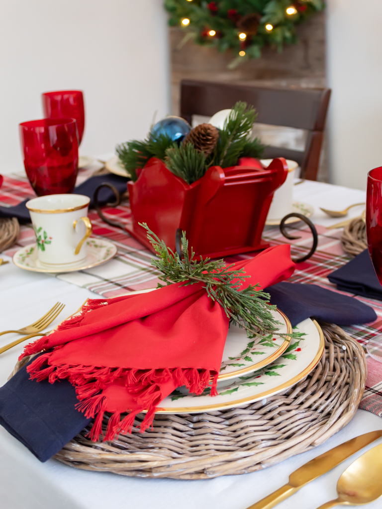 Dining room table set for Christmas with red and blue decor, Lenox Christmas china, a plaid table runner, and a red sleigh centerpiece filled with ornaments and greenery