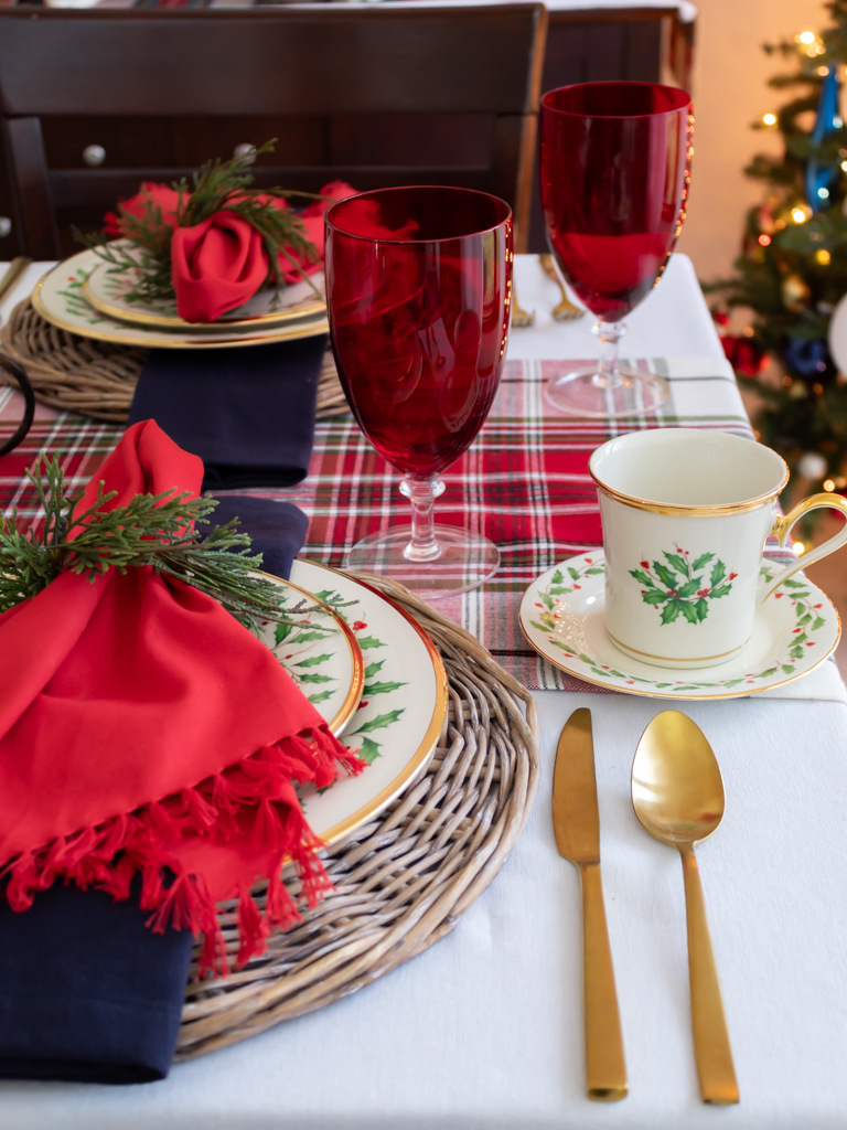 Christmas place setting on a holiday table with Lenox Christmas china on a woven charger with red and blue fabric napkins with a greenery napkin ring, red goblets, gold silverware, and a plaid table runner