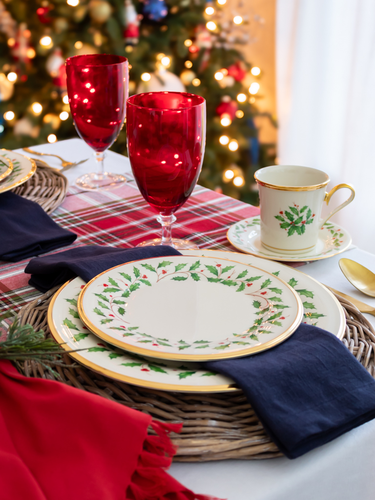 Christmas place setting on a holiday table with Lenox Christmas china on a woven charger with red and blue fabric napkins, red goblets, gold silverware, and a Christmas tree in the background