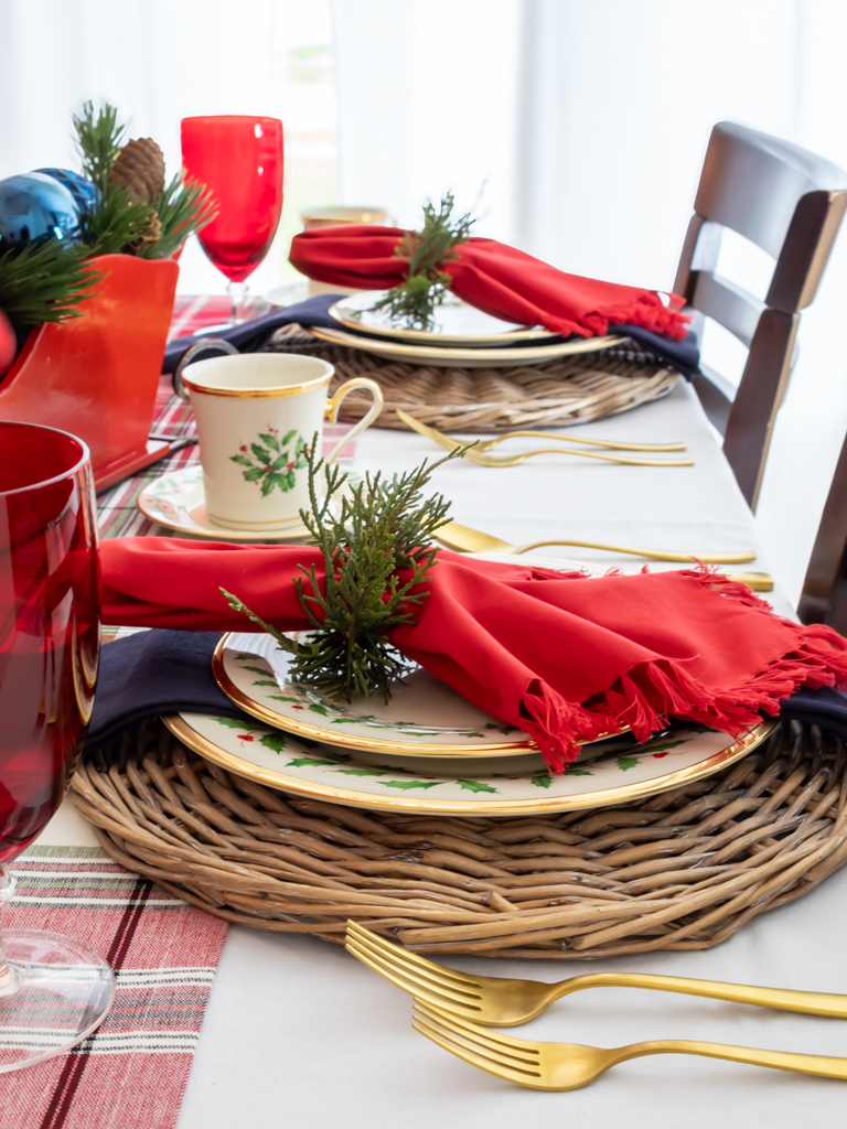 Christmas place settings on a holiday table with Lenox Christmas china on a woven charger with red and blue fabric napkins with a greenery napkin ring, red goblets, gold silverware, and a plaid table runner