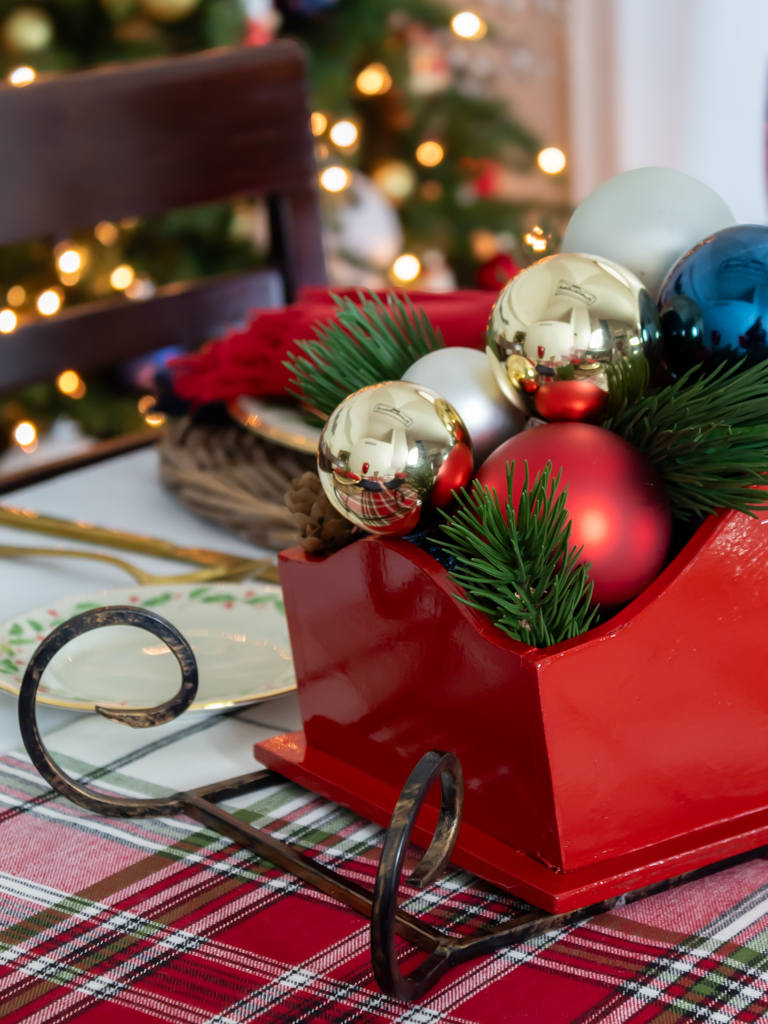 Close up view of DIY red Santa sleigh Christmas centerpiece set on a holiday tablescape filled with red, gold, white and blue ornaments and greenery