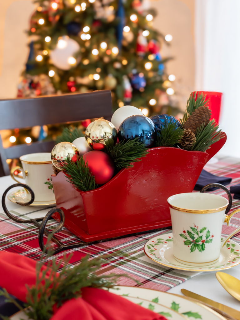 DIY red Santa sleigh Christmas centerpiece set on a holiday tablescape filled with red, gold, white and blue ornaments and greenery and a Christmas tree lit up in the background