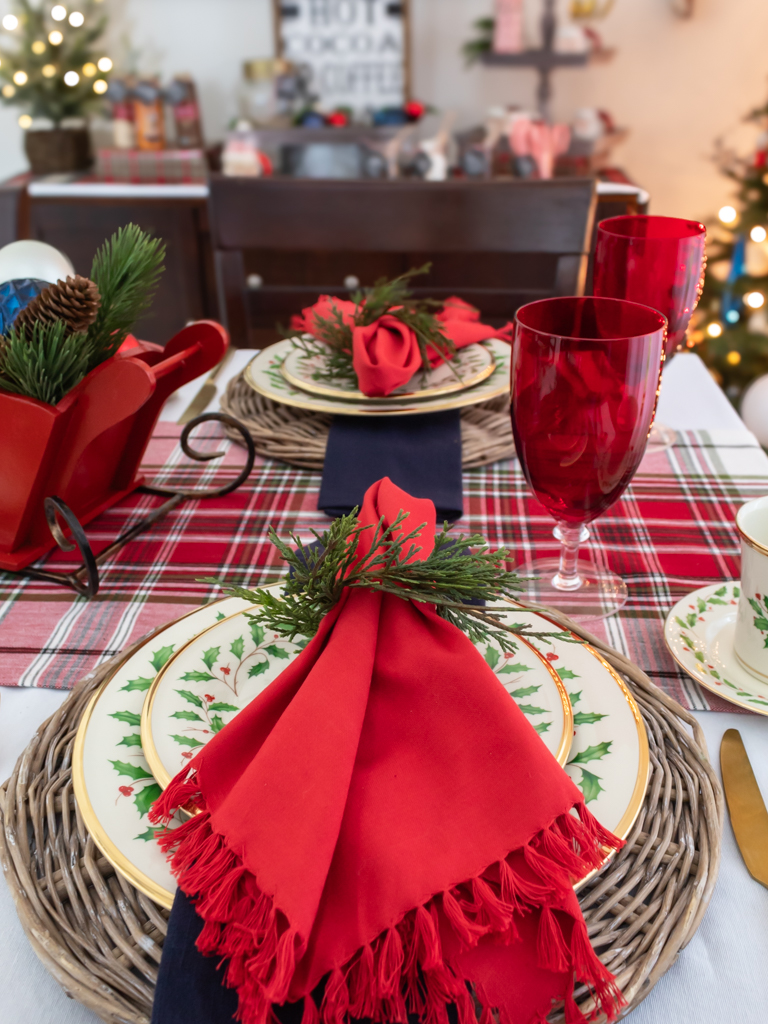 Dining room table set for Christmas with red and blue decor, Lenox Christmas china, a plaid table runner, a red goblet, and a red sleigh centerpiece filled with ornaments and greenery with a hot cocoa buffet in the background