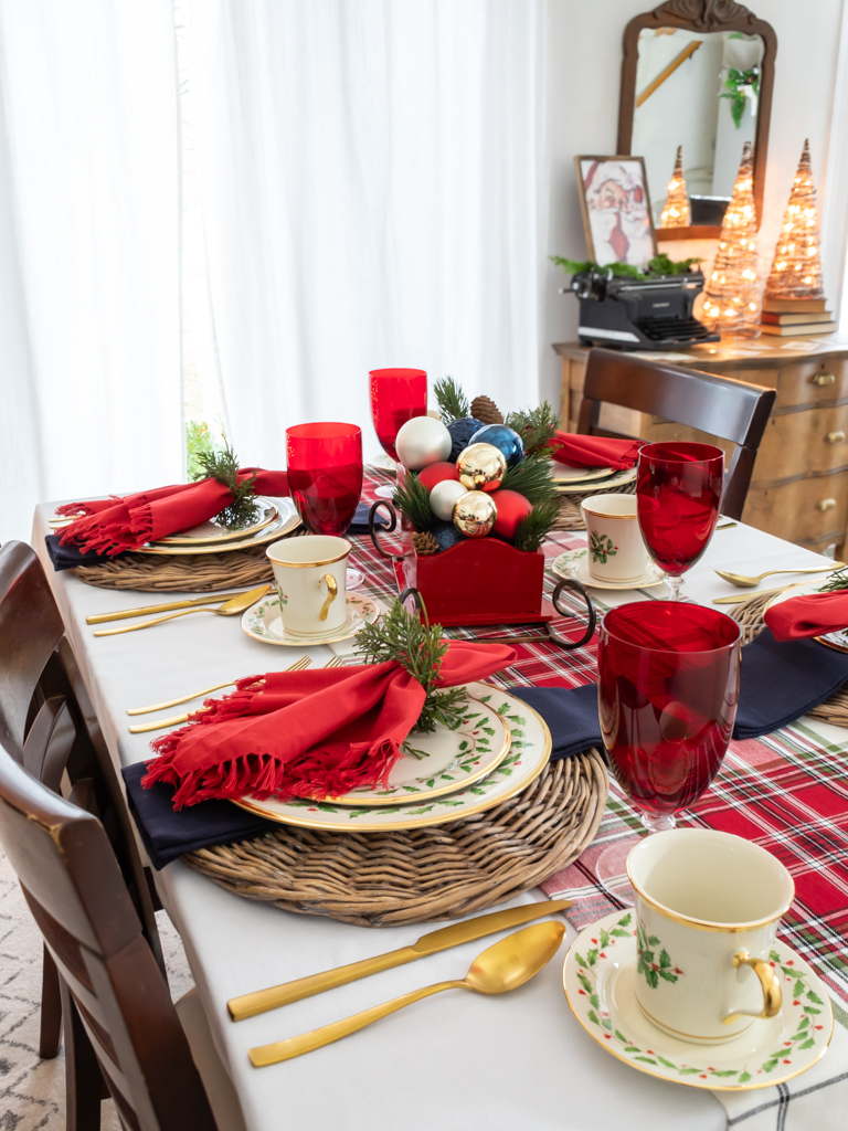 A tablescape set for a holiday meal, with red and blue decorations including Lenox Christmas china, a red fringed napkingwith a greenery napkin ring, red goblets, a red sleigh centerpiece filled with ornaments and a dresser and mirror in the background with mini trees and a framed santa print