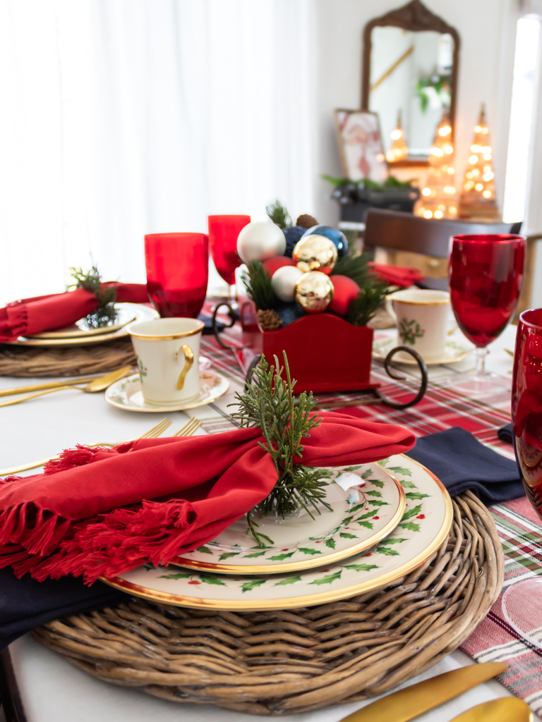 Dining Room table decorated for Christmas with holiday dishes, read and blue decor, and Santa sleigh centerpiece filled with ornaments and greenery. 