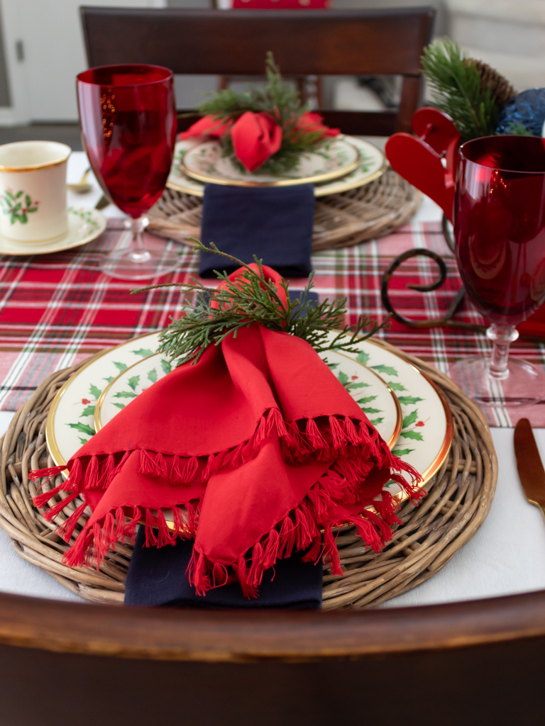 Christmas place setting on a holiday table with Lenox Christmas china on a woven charger with red and blue fabric napkins with a greenery napkin ring, red goblets, gold silverware, and a plaid table runner