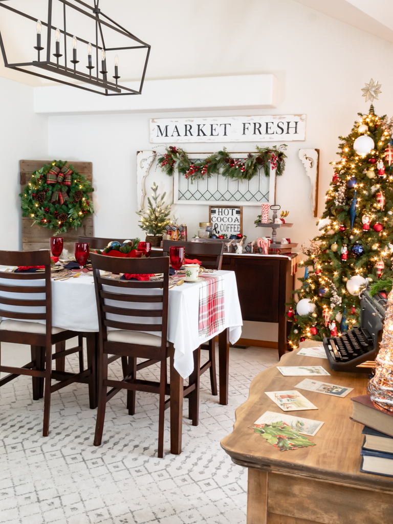 Dining room decorated for Christmas with red and blue decor, a tablescape set for a holiday meal, and a cozy hot cocoa bar set on a buffet in the background and a antique dresser with a holiday vignette in the foreground